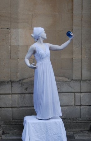 A washer-woman style white living statue who performs highly skilled contact juggling with her aqua blue magical water droplet.